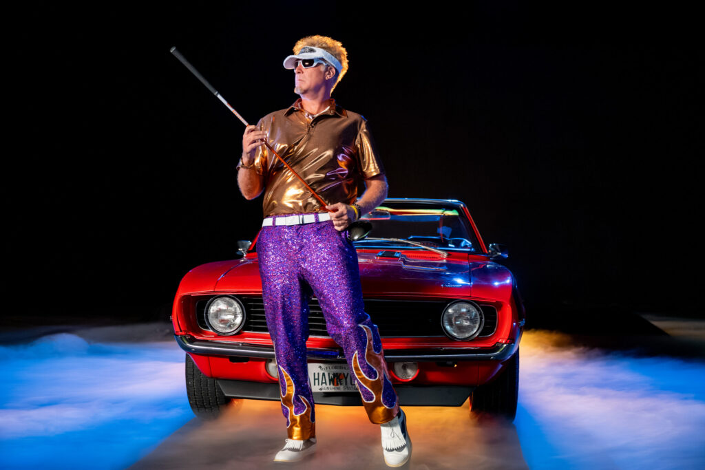"Man in shiny shirt, purple sequin pants, and white sunglasses holding a cane, standing in front of a red vintage convertible car with mist and colorful lighting, black background, dramatic and retro style scene."